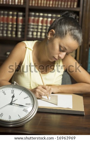 Woman seated at desk writing with clock on desk. Vertically framed photo.