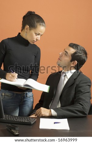 Woman takes notes for a business man sitting at a desk. Vertically framed photo.