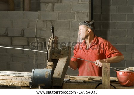The worker has a piece of metallic bar.  He is cutting it down to the size he needs.  Horizontally framed shot.