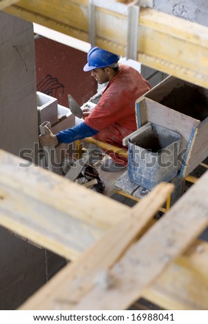 The worker is crouched on a platform.  He is assembling the cinder rock wall.  Vertically framed shot.