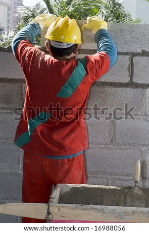 The worker is standing in front of the wall.  He is assembling more cinder blocks to the wall.  Vertically framed shot.