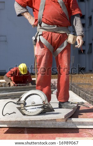 The worker is standing behind a circular saw.  There is another worker in the background.  Vertically framed shot.