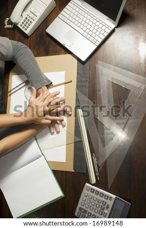 Two hands on desk surrounded by drafting tools, laptop, phone, and calculator. Vertically framed photo.