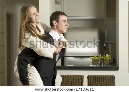 Young, smiling couple stand in kitchen as he gives her a piggyback ride. Horizontally framed photo.