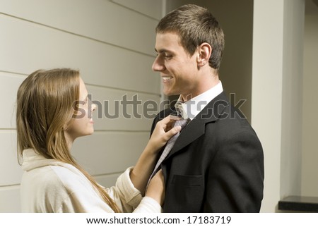Young couple smiling as she fixes his tie, he is in a suit and she is in a bathrobe. Horizontally framed photo.