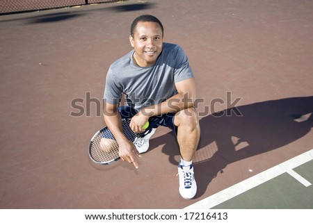 Smiling tennis player kneeling down on the tennis court holding his racket and ball. Horizontally framed photo.