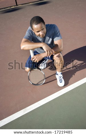 Tennis player crouching down looking defeated and sad, he holds his tennis racket and rests his arms on his knees. Vertically framed photo.