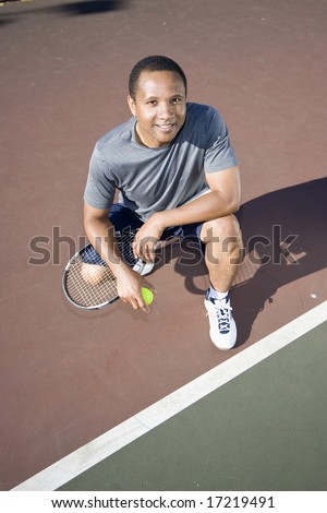 Smiling tennis player kneeling down on the tennis court holding his racket and ball. Vertically framed photo.