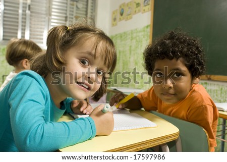 Children sitting at desks in a classroom.  They are working together and are facing the camera.  Horizontally framed shot.