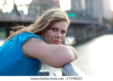 Young woman leaning against a bridge with buildings in the background smiling a sly smile. Horizontally framed photo.