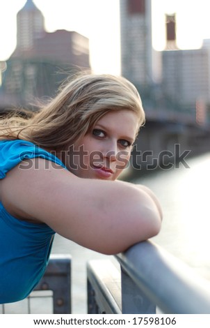 Young woman leaning against a bridge with buildings in the background smiling a sly smile. Vertically framed photo.