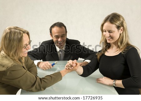 Two women and a man at a table smiling, and the women are arm wrestling. Horizontally framed photo.