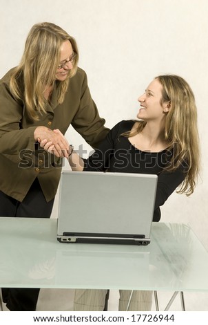 Two people are sitting together in a room at a table. The two woman are shaking hands and smiling at each other.