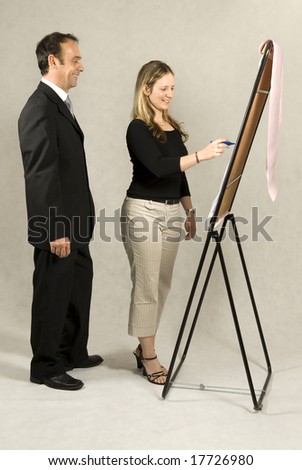 A man is standing next to a young woman who is drawing on a large board.  It looks as if she is drawing a business model.  They are smiling and looking at the board.  Vertically framed shot.
