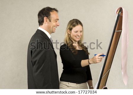 A man is standing next to a young woman who is drawing on a large board.  It looks as if she is drawing a business model.  They are smiling and looking at the board.  Horizontally framed shot.