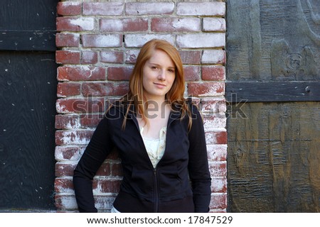 Attractive girl poses for camera by leaning against a brick wall. She is smiling at the camera. Horizontally framed photo