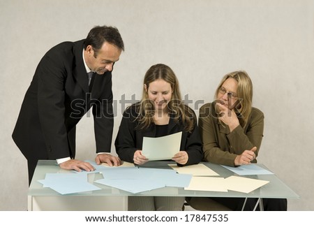 Two women at desk looking at paperwork while a man stands beside them. Horizontally framed photo.