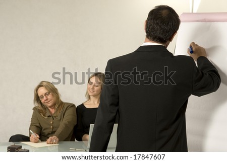 A Boss in instructing his employees in a business meeting.  He is writing on a large presentation board.  The older woman is taking notes and the younger woman is looking down at the paper.