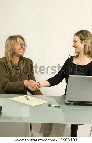 Two Women are sitting at a table in a business meeting.  They are shaking hands and smiling at each other.  Vertically framed shot.