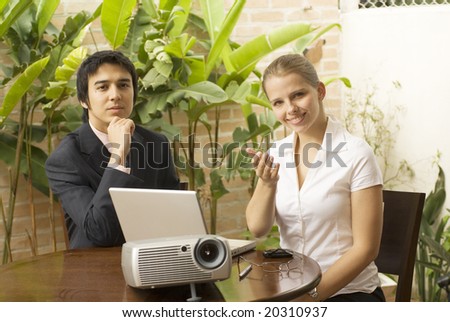 Man and woman with a projector and a laptop. They are seated and she is smiling while he looks pensive. Horizontally framed photo.