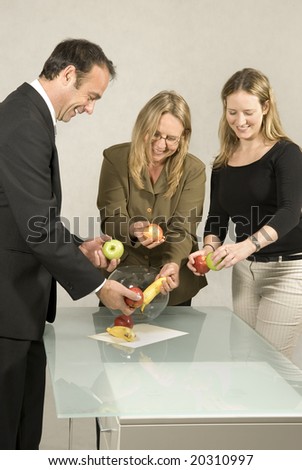 Three people are in an office.  It appears that they are about to arrange the fruit into the glass bowl.  Vertically framed shot.