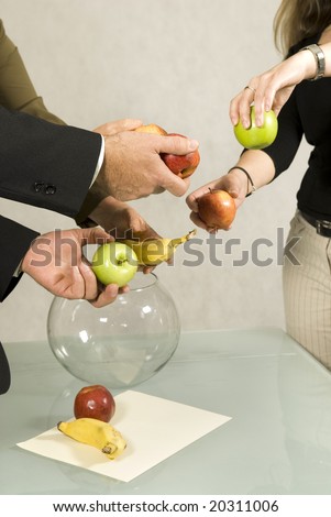 Three people are in an office.  It appears that they are about to arrange the fruit into the glass bowl.  Vertically framed shot.