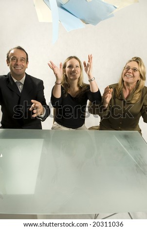 Three people are in an office in a meeting.  They are smiling and throwing papers in the air in success.  Vertically framed shot.