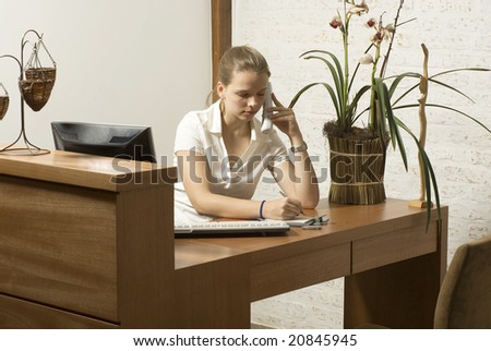 Woman on the phone sitting at a desk with a computer and a plant. Horizontally framed photo.