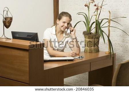 Smiling young woman sitting at a desk talking on the phone.  There is a computer and a plant. Horizontally framed photo.