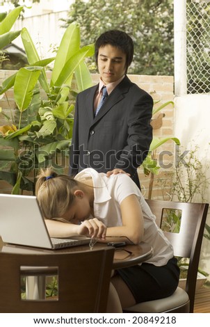Man and woman at a desk with a laptop computer. He is comforting her as she lays her head on the table. Vertically framed photo.