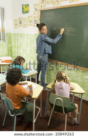 Elementary school teacher standing at a chalkboard writing as she smiles at her students writing at their desks. Vertically framed photo.