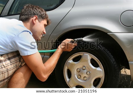 A man washing his car.  He is using a hose to wash the tires. Horizontally framed shot.