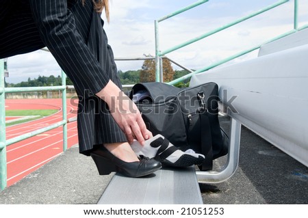 Woman in a suit sitting on a bleacher at a track putting on a running shoe. Horizontally framed photo.