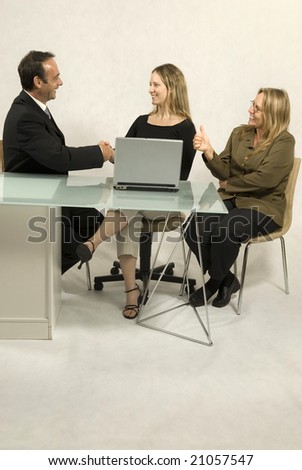 Two women and a man seated at a desk in front of a laptop, they are all smiling and the man and one woman are shaking hands.