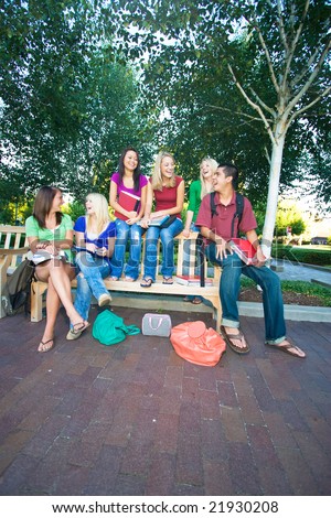 Group of five laughing high school girls and one boy sitting on a bench holding books. Vertically framed photo.