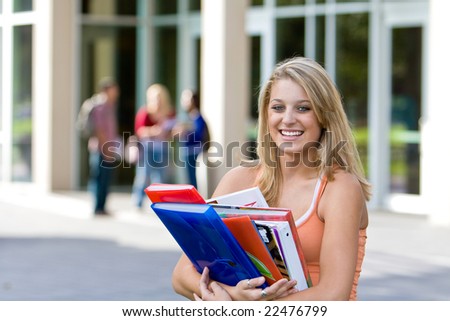 Young smiling female student carrying her books outside of school.  There are kids in the background. Horizontally framed photo.