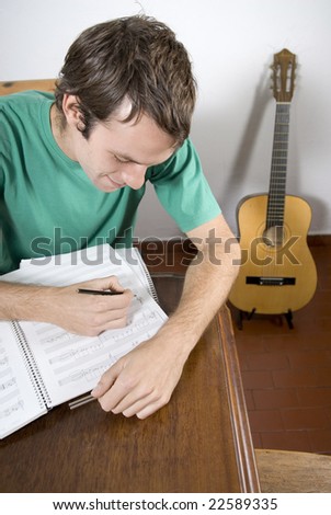 Man seated at a desk writing music with a guitar in the background. Vertically framed photo.