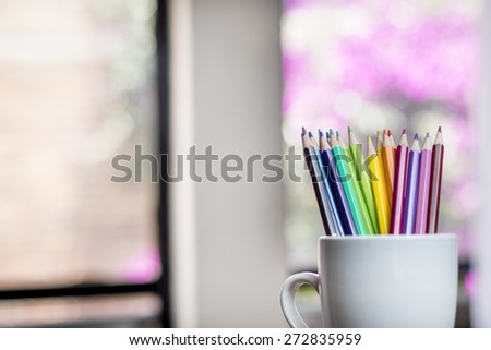 A group of color pencils in shadows in a silhouette cup with a blurred window and garden background