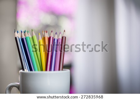 A group of color pencils in shadows in a silhouette cup with a blurred window and garden background