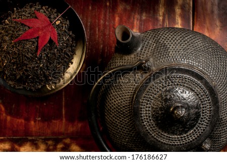 Japanese iron teapot and heap of tea leaves from top. Red mable leaf on top of tea leaves. Mahogany background. Asian culture background. Horizontal photography no people.