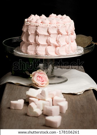 Scalloped pale pink cake on glass stand next to rose and marshmallows on wooden table. Still live photo, no people.