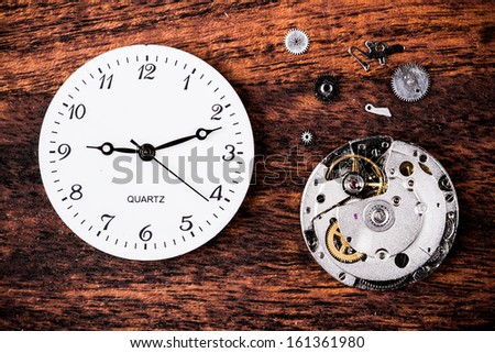 Various clock parts (cogs, hands, springs) laid on a rustic/antique wood background, viewed from above