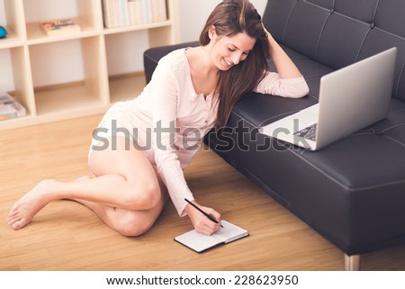Smiling young woman is writing on your notebook while is working with her laptop sitting on floor at home