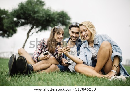 Group of friends two women and one man using a smartphone, sitting on floor in the park