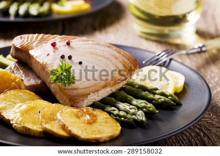 Grilled tuna steak served on asparagus with roasted potatoes on a black plate.
