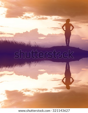 Silhouette of woman standing alone at the field during beautiful sunset with water reflection