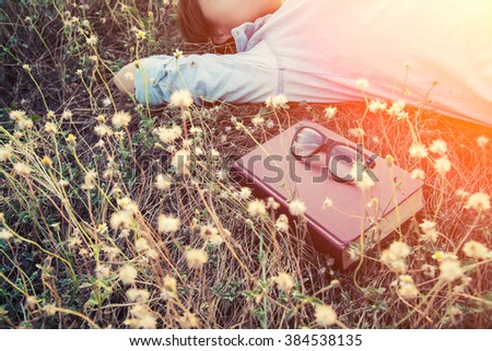 young woman napping on the dandelion field after reading book put out glasses on the book