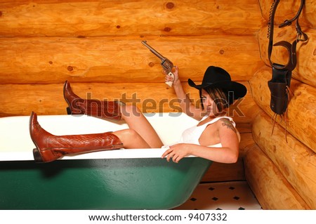 cowgirl in cowboy boots and hat in claw-foot bath tub holding silver pearl handled revolver with holster hanging on wall in log cabin