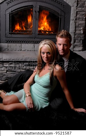 romantic couple sitting together in front of roaring fire in stone fireplace in suit and tie and dress