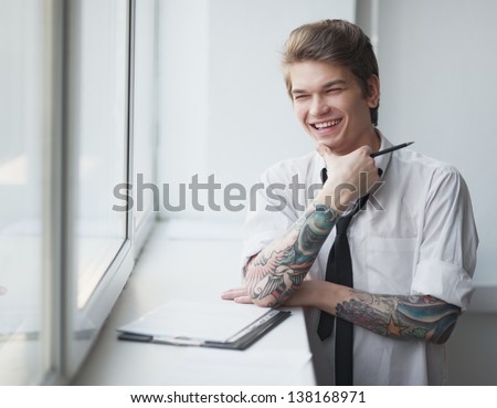 the beautiful young man in a shirt with a tattoo writes at a window to studios